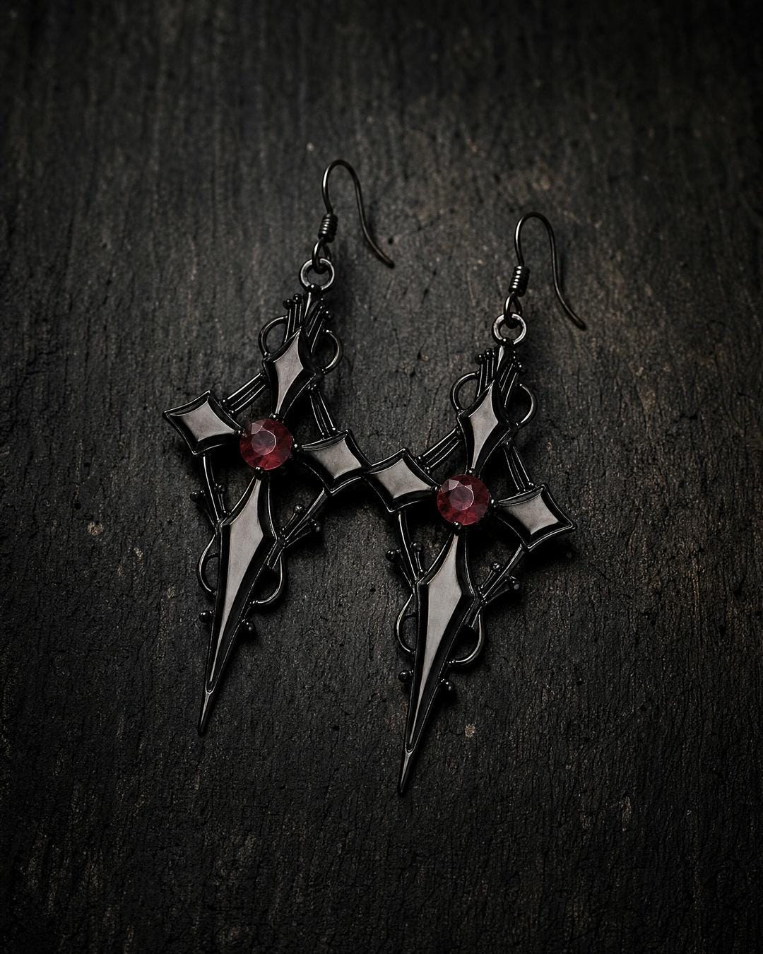 Pair of black cross dagger earrings with a red faceted stone and tapered spike, shown flat on a surface.