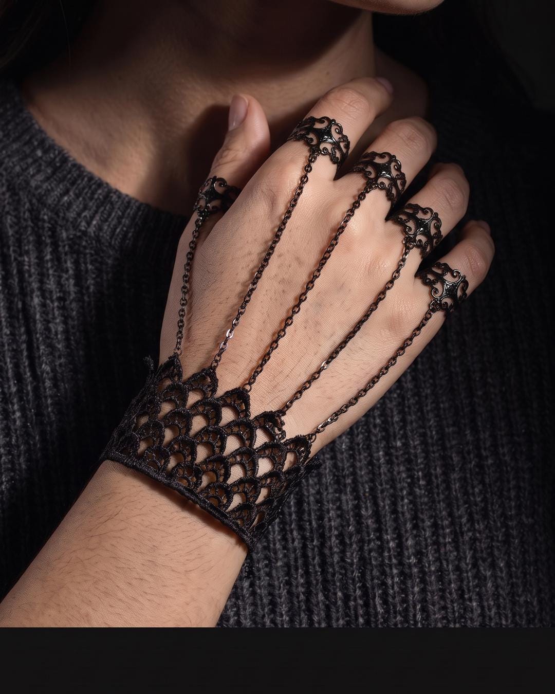 Lace hand jewelry worn on a hand against a dark background, chains crossing to four filigree rings.