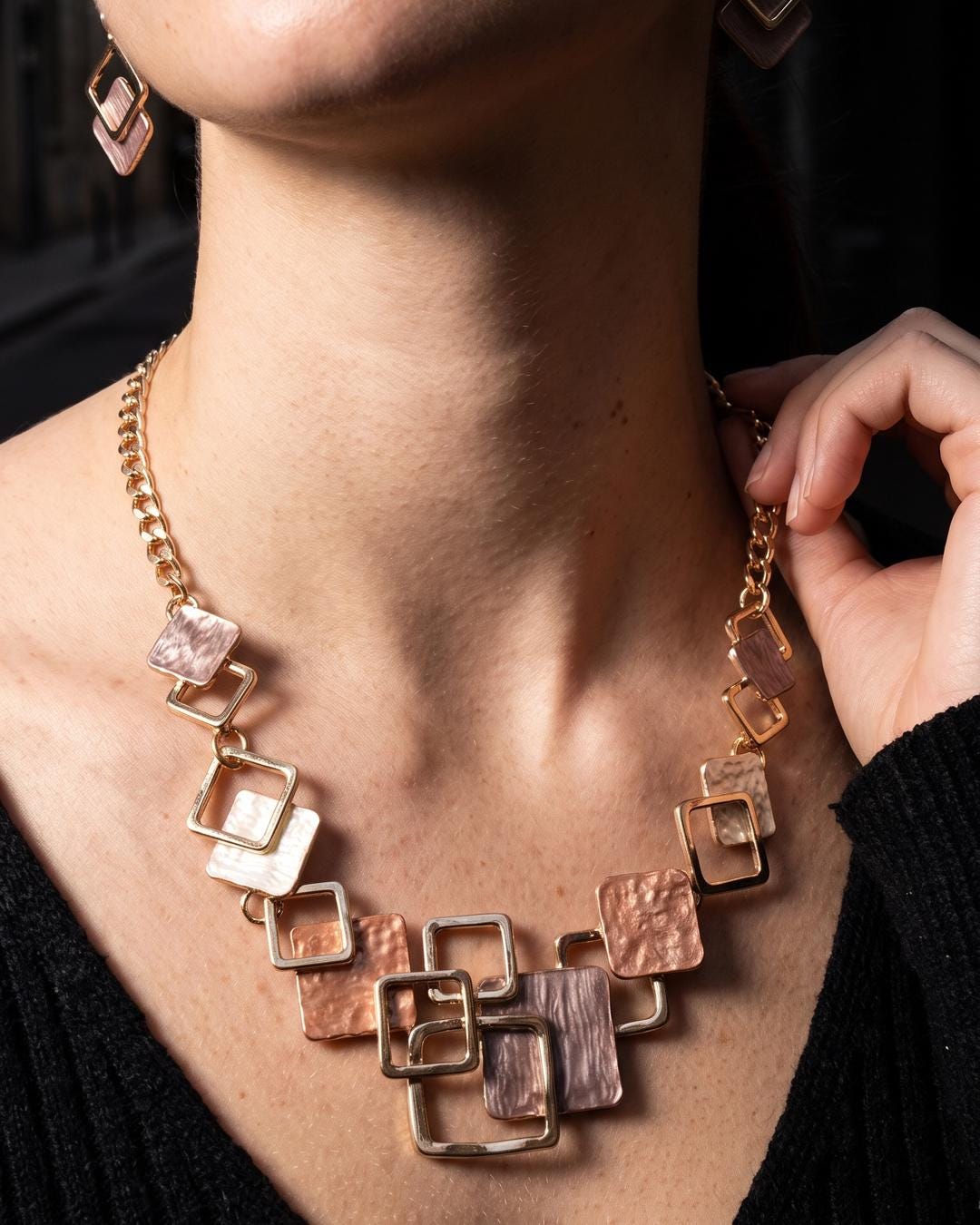 Geometric necklace worn at the throat over a dark knit, square frames catching warm light